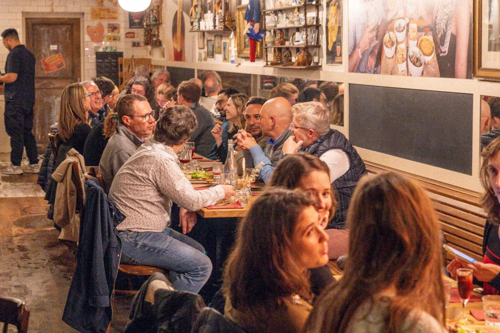 Une longue tablée d'estaminet en plein service, convives serrés, ardoises au mur.